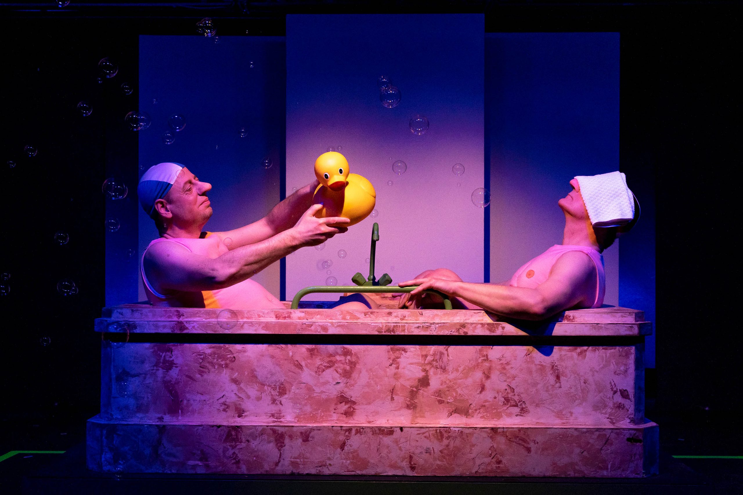 Zwei Männer mit Badekappe sitzen sich in einer Badewanne gegenüber.