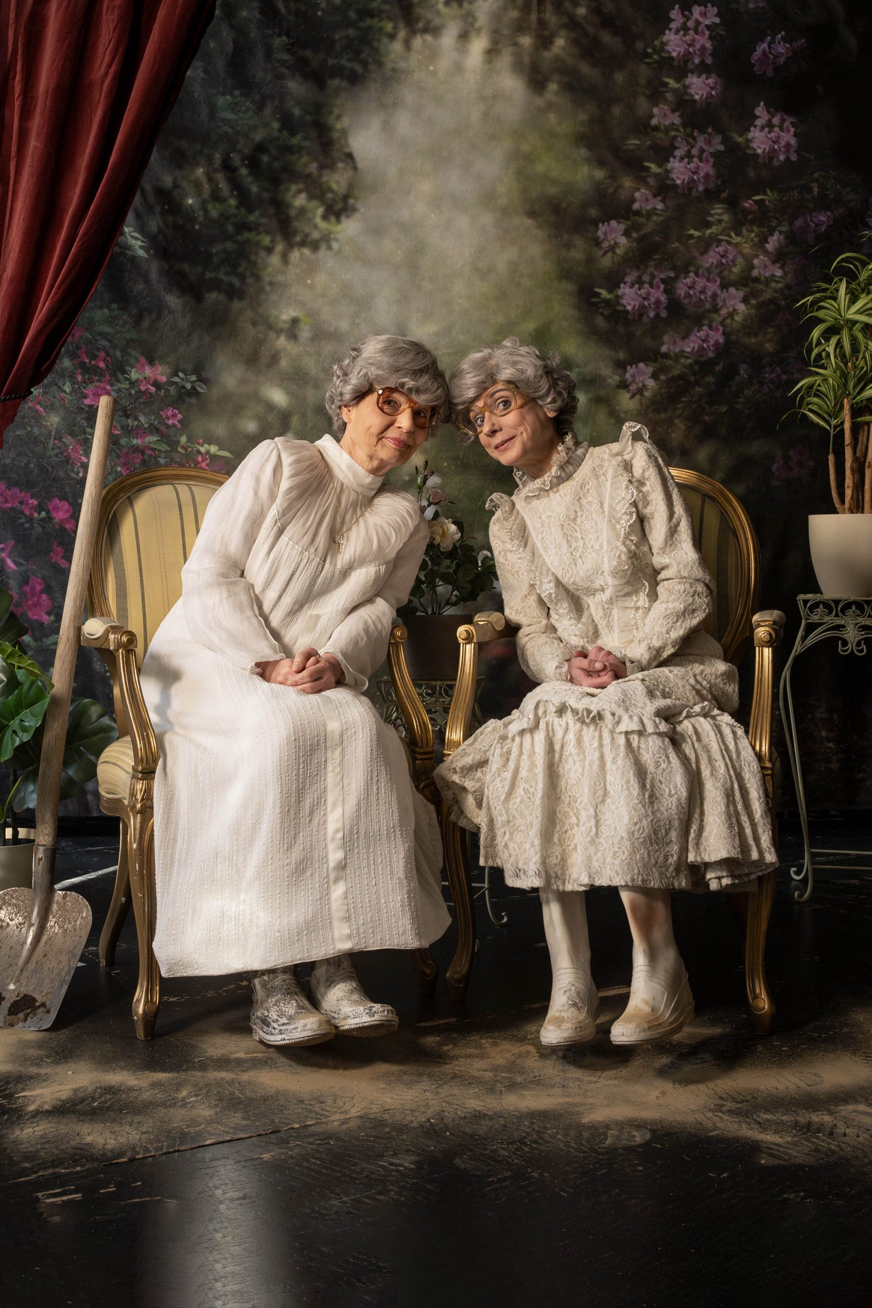 Martha und Abby sitzen ganz in Weiß in einer Art Fotostudio auf zwei altmodischen Stühlen, an denen eine dreckige Schaufel lehnt.